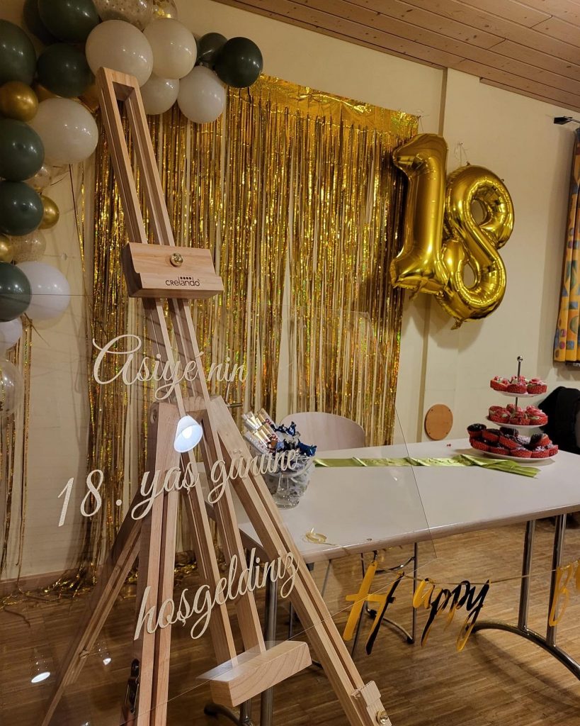 Geburtstags Hintergrund mit goldenen fäden die herrunter hängen rechts daneben eine goldene 18 in einem folienballon der mit helium schwebt. links davon eine ballon girlande und vorne dran ein stativ mit einem acrylglas länglich wo drauf steht asiyenin 18. yas gününe hosgeldin.