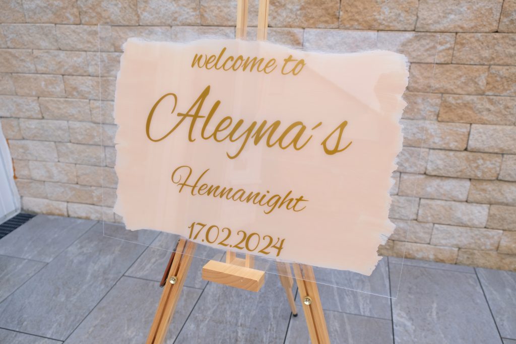 Auf einem Stativ stehendes Acrylglas mit beigem Hintergrund. Vorne ddrauf steht in geschwungener Schrift:" welcome to Aleynas Henna night 17.02.2024"
