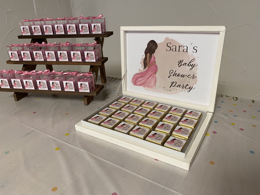 links eine etagerie aus treppen mit acryl boxen klein ( 4x4cm ) mit rosa kerzen und vorne ein bild mit einer schwangeren frau und einer schrift " Sara´s Baby Shower Party ) rechts daneben eine Pralinen Box mit VIereckigen schokoladentäfelchen mit dem gleichen Bild der Schwangeren und der Schrift verziert und oben an der klappe der Pralinen Schachtel auch das gleiche Bild.