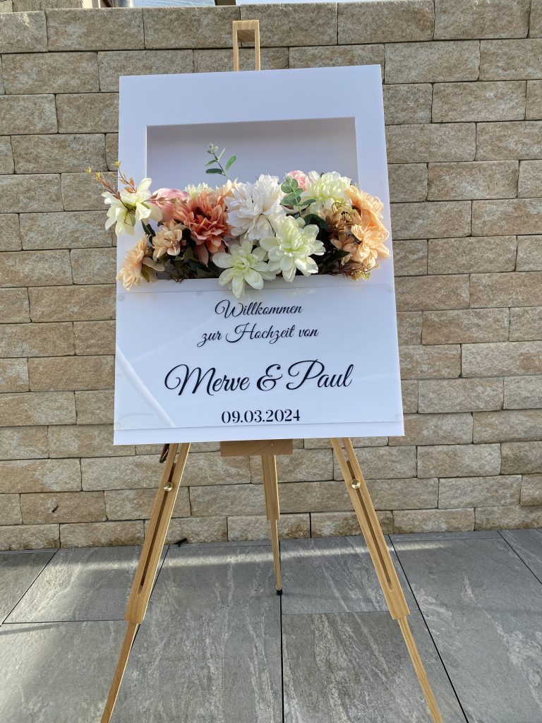 Auf einem Stativ stehendes in 3D aussehende Polaroid Box mit blumen im aussschnitt. unten drunter steht willkommen zur hochzeit von merve und Paul